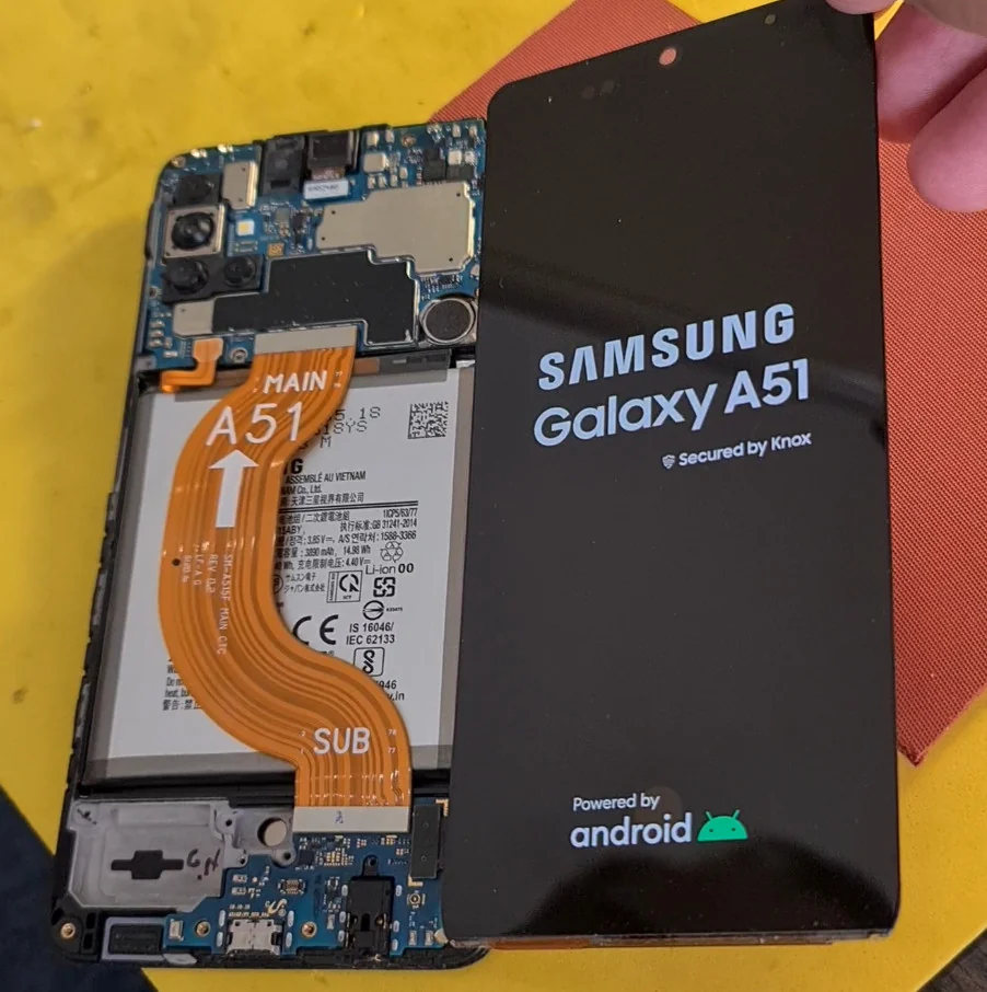 Разобранный Samsung Galaxy A51 на жёлтом столе: видна материнская плата, аккумулятор и оранжевый межплатный шлейф с маркировкой MAIN A51, справа лежит работающий дисплейный модуль с заставкой Samsung