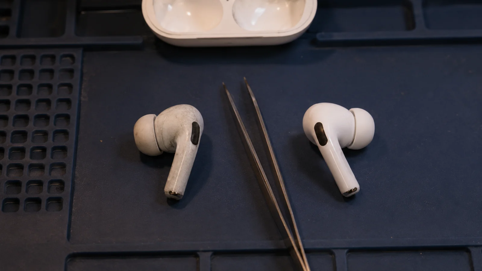 Два белых наушника AirPods лежат рядом на тёмном рабочем коврике: один потёртый и пожелтевший, второй новый, между ними пинцет