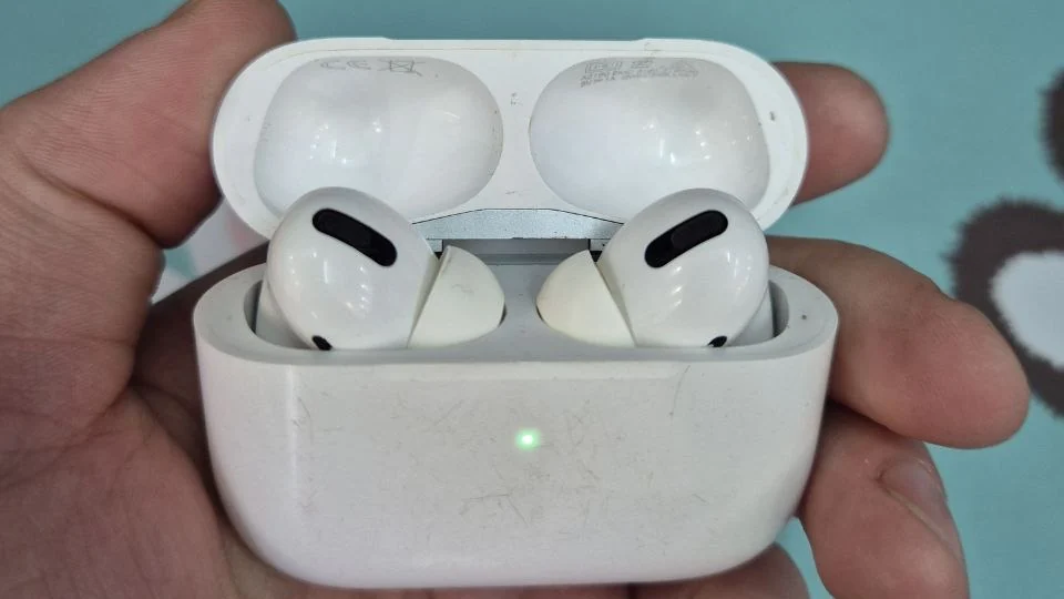 Открытый кейс AirPods Pro на ладони, оба наушника внутри, корпус кейса в мелких царапинах, на передней стенке горит зелёный индикатор