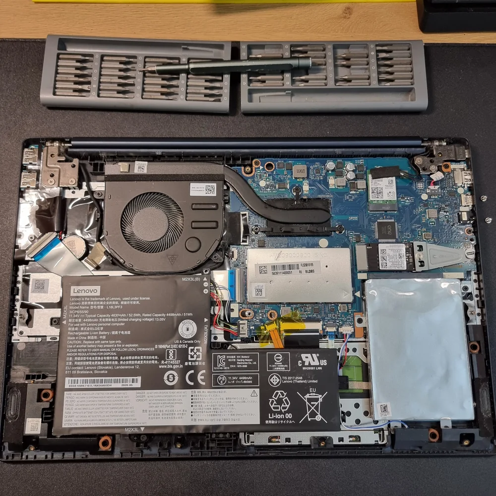 Ноутбук Lenovo со снятой нижней крышкой на столе мастерской: слева аккумулятор с шильдиком L18L3PF3, в центре вентилятор и теплотрубка, справа накопитель M.2 и жёсткий диск, сверху лежит набор бит