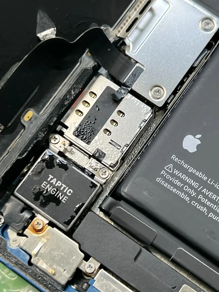 Вскрытый корпус iPhone: справа родная батарея Apple с предупреждающей надписью на корпусе, слева Taptic Engine и лоток SIM-карты, на металле видны следы жидкости