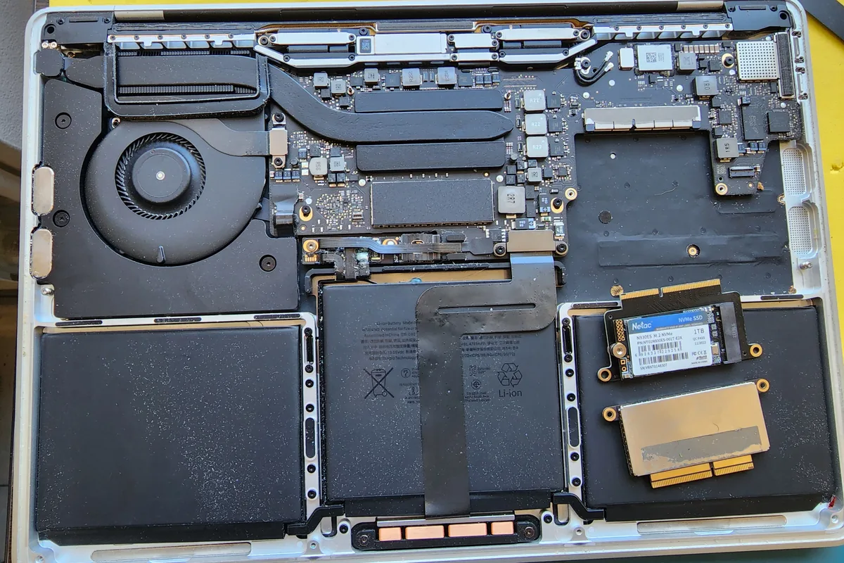 MacBook Pro на столе мастера: нижняя крышка снята, видны секции аккумулятора, материнская плата и система охлаждения