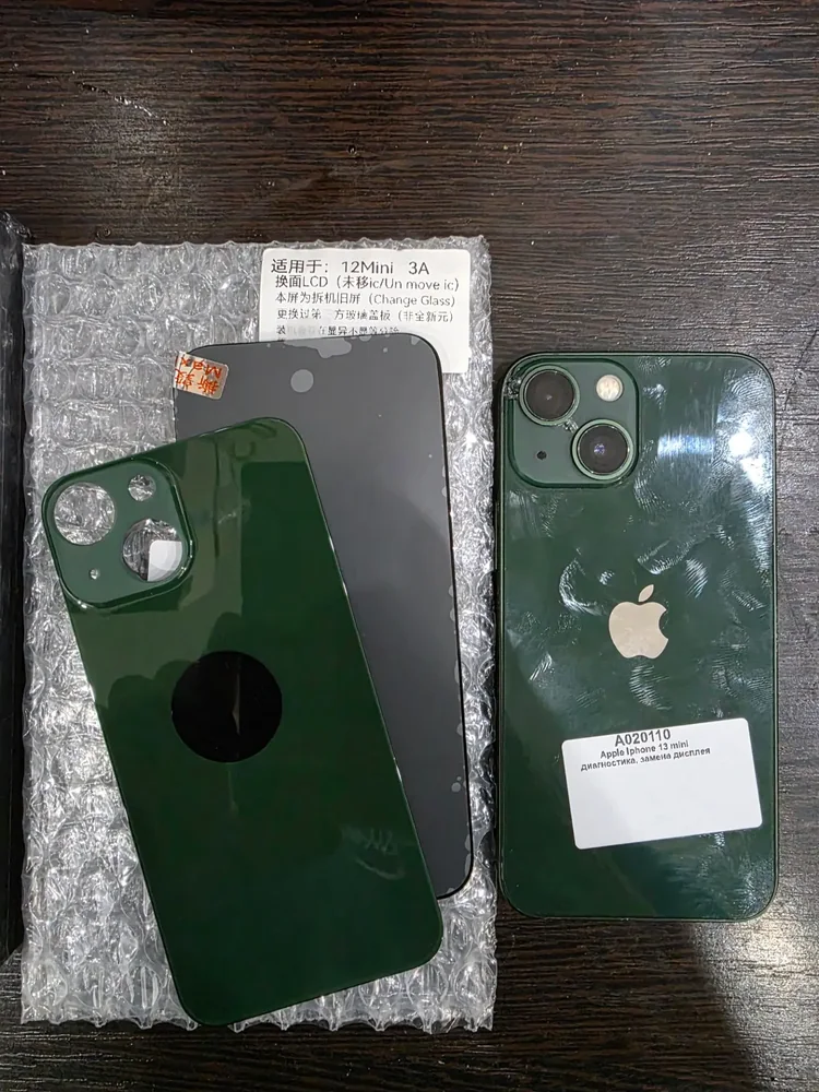 Заказ на замену дисплея iPhone 13 mini в мастерской: аппарат с биркой заказа, рядом новый модуль экрана в упаковке и запасная задняя крышка