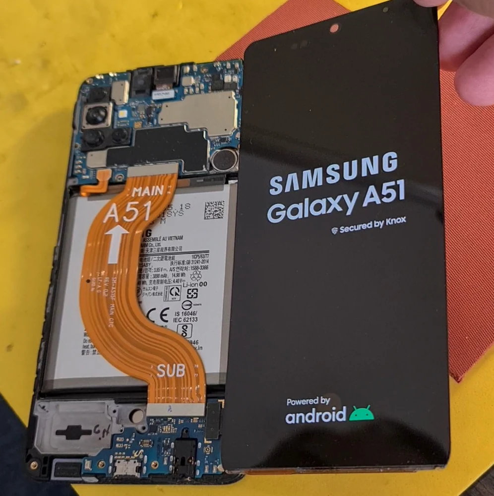 Разобранный Samsung Galaxy A51 на рабочем столе мастера: видны материнская плата, аккумулятор и шлейфы, рядом лежит подключённый рабочий дисплей с заставкой