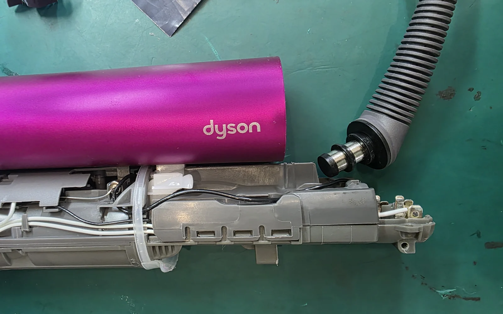 Розовая рукоятка Dyson со снятым корпусом лежит на зелёном коврике: под корпусом серая внутренняя рама с проводами, стянутыми хомутом, справа отсоединён гофрированный шланг.