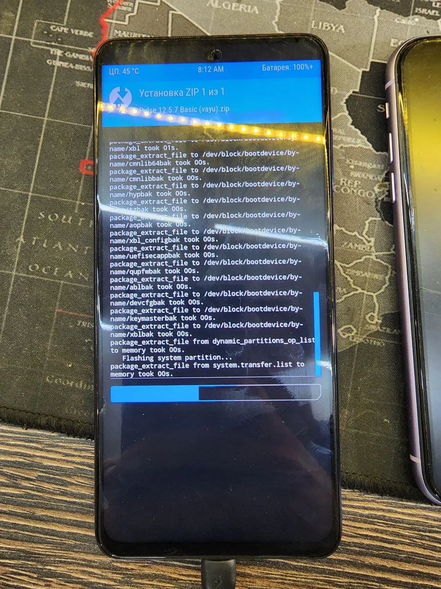 Смартфон Poco X3 Pro лежит на ремонтном коврике с картой мира, на экране идёт установка прошивки, строки лога и полоса прогресса