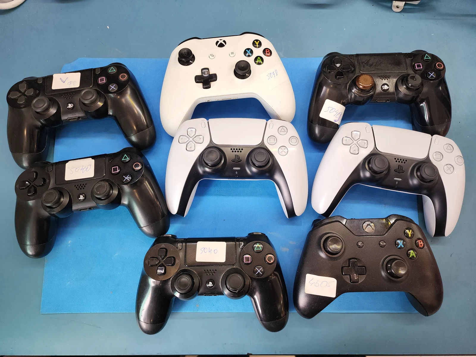 Семь геймпадов от PlayStation 4, PlayStation 5 и Xbox лежат рядом на синем ремонтном коврике мастерской с приёмными бирками
