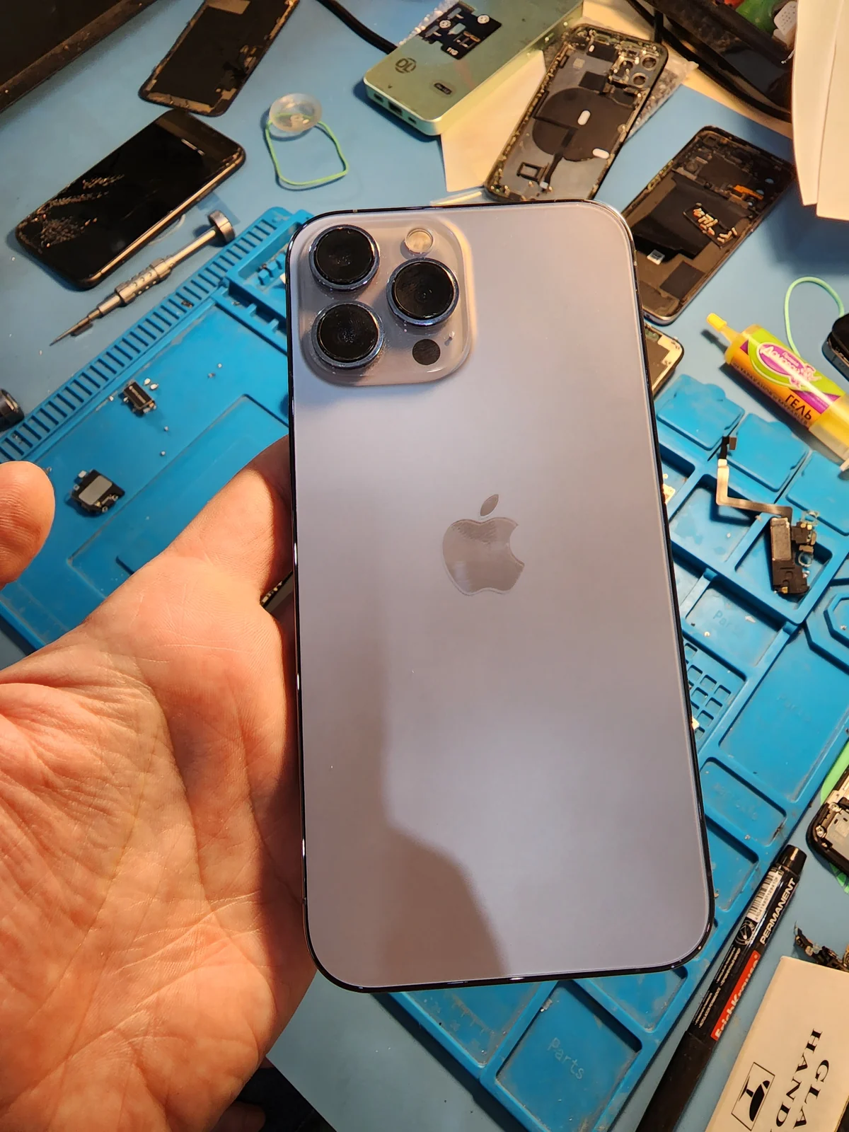 Сиреневый iPhone 13 Pro Max задней стороной вверх лежит на ладони мастера, за ним синий рабочий коврик с разобранными аппаратами и инструментом