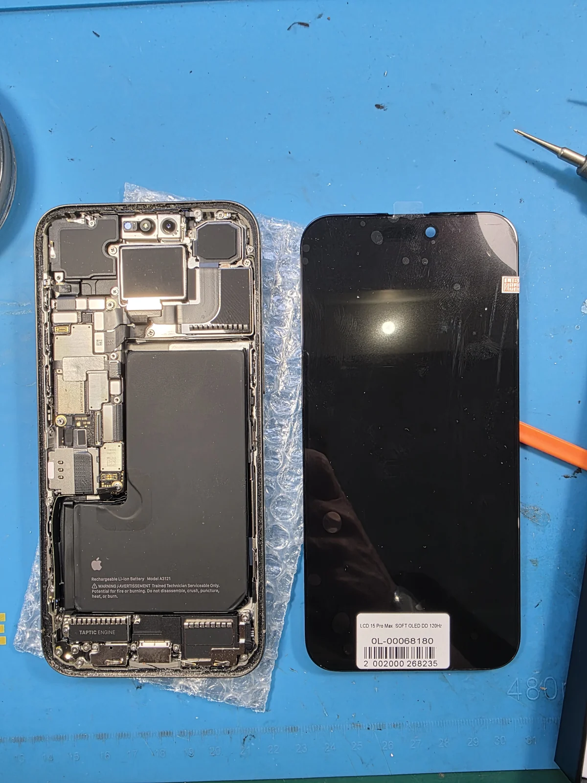 Вскрытый корпус iPhone 15 Pro Max с платой, аккумулятором и Taptic Engine, рядом новый дисплейный модуль с этикеткой поставщика на синем коврике мастерской