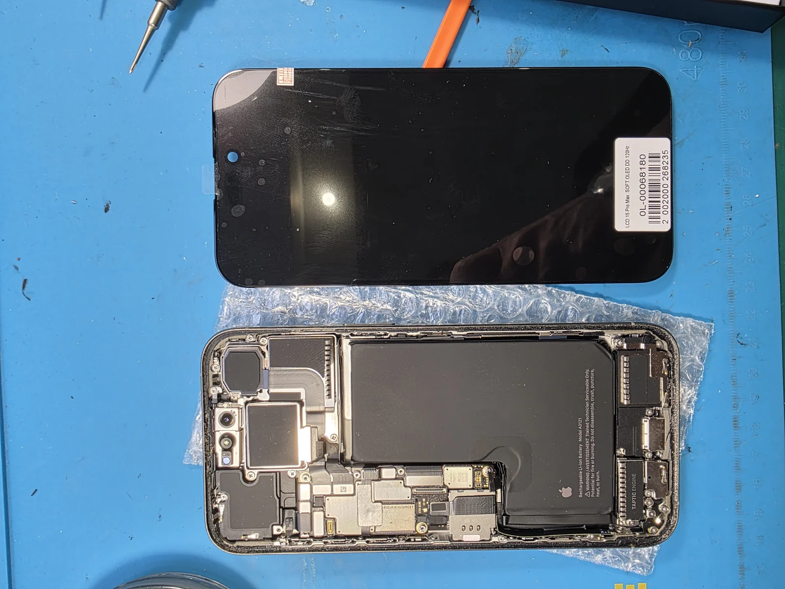 Новый дисплейный модуль iPhone с этикеткой поставщика, где указан тип матрицы и частота обновления, ниже на коврике лежит вскрытый корпус телефона с платой и аккумулятором