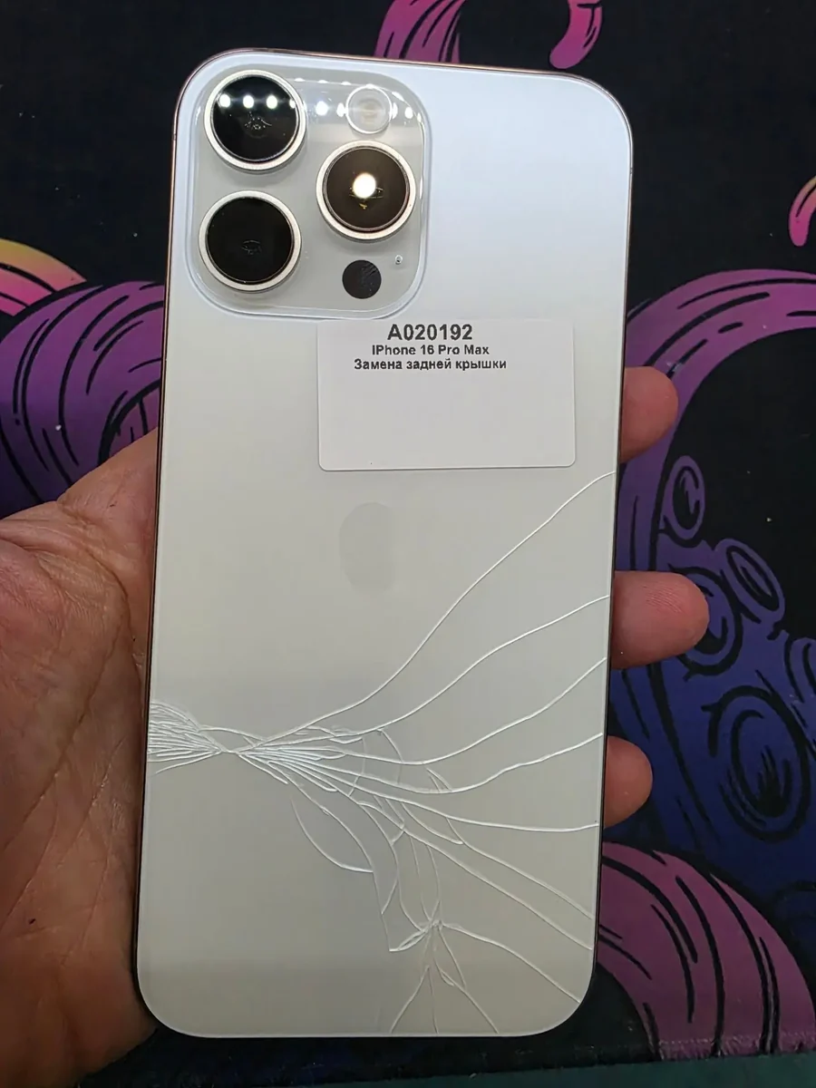 Белый iPhone 16 Pro Max в руке мастера задней стороной вверх: заднее стекло треснуло лучами от нижнего угла, сверху приклеена наклейка приёмки с номером заказа