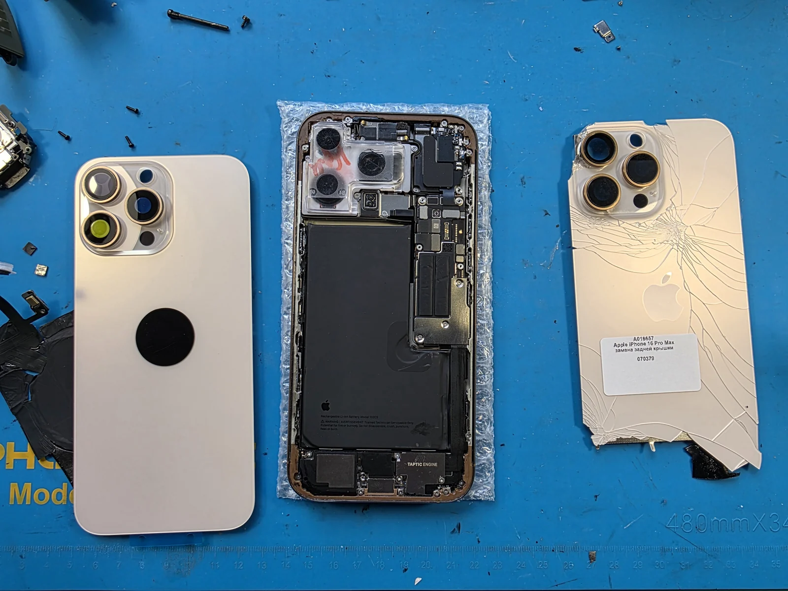 Разобранный iPhone 16 Pro Max на синем коврике: слева новая задняя крышка, в центре корпус с платой, аккумулятором и Taptic Engine, справа старая разбитая крышка с наклейкой приёмки