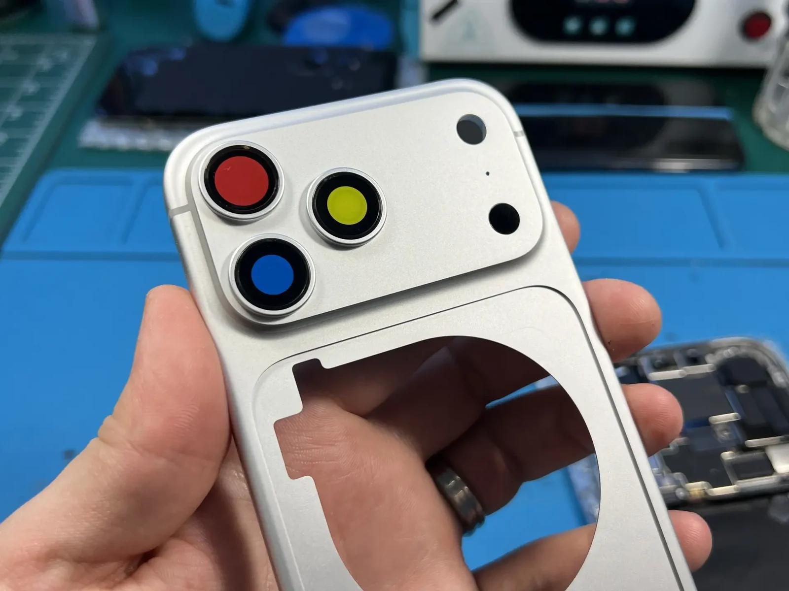 Корпус iPhone 17 Pro серебристого цвета в руке мастера: камерная площадка во всю ширину, три объектива под цветной защитной плёнкой, вырез под MagSafe