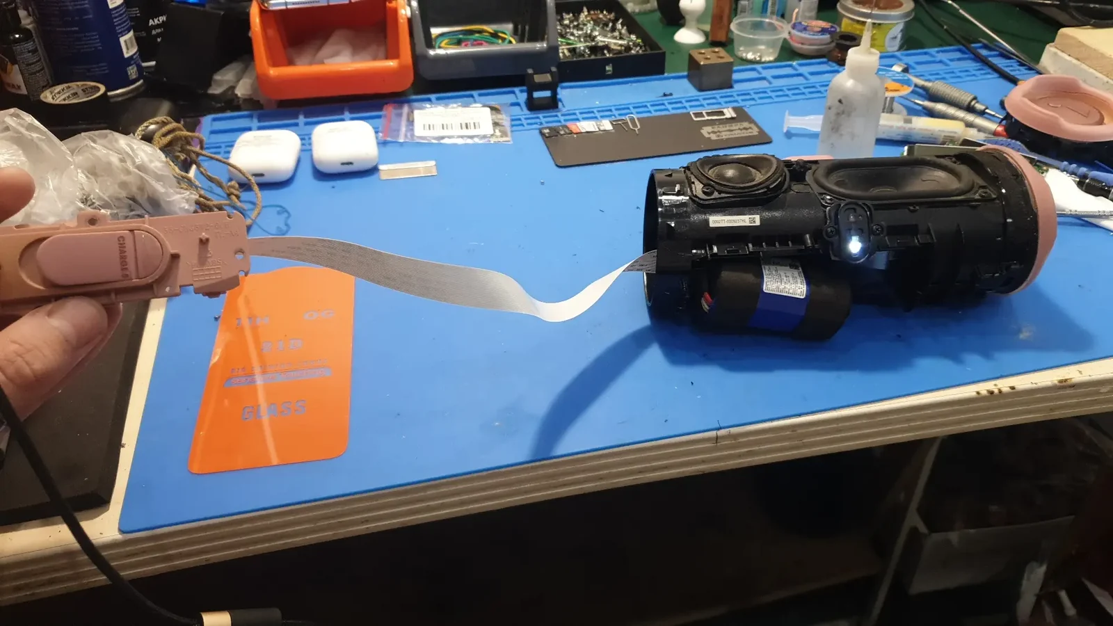 Вскрытая колонка JBL Charge лежит на столе, в руке мастера панель кнопок, от которой к корпусу тянется длинный светлый шлейф, рядом виден синий аккумулятор.