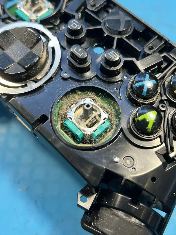 Разобранный геймпад Xbox крупным планом: колпачок стика снят, вокруг его модуля на зелёной плате скопились волосы и пыль.