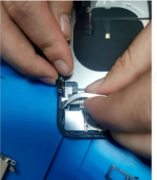 Замена кнопки на iPhone X у нас в мастерской: руки мастера пинцетом заводят белый шлейф кнопок в разобранный корпус, лежащий на синем коврике