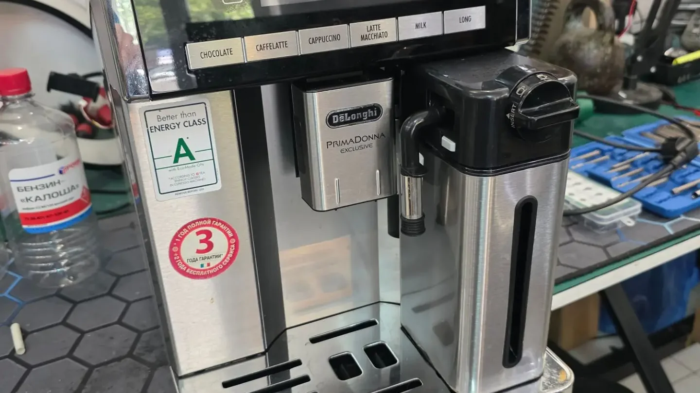 Кофемашина DeLonghi Primadonna Exclusive стоит на приёмном столе мастерской, на фоне полки с инструментом и катушками провода