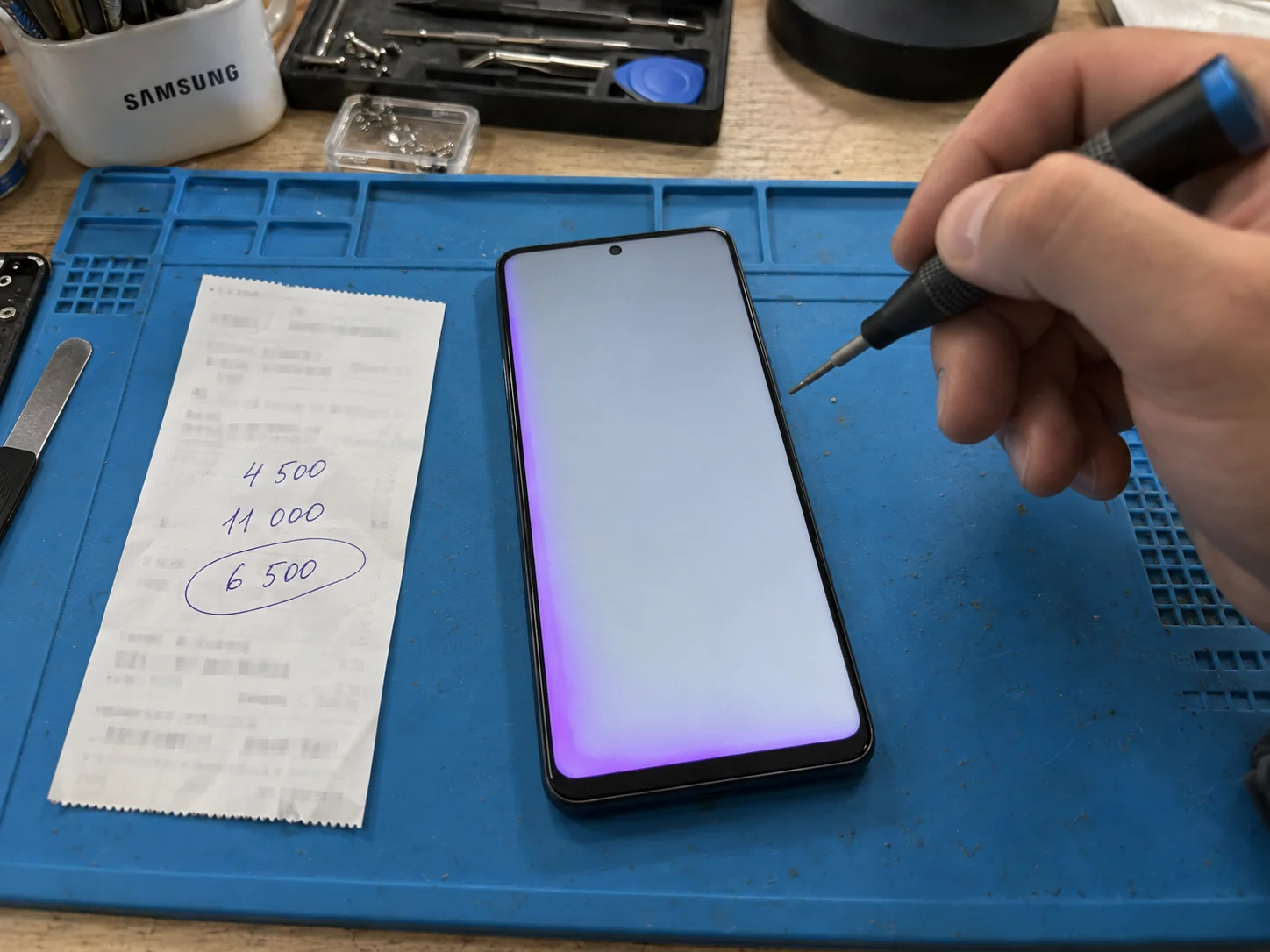 Samsung с фиолетовым отливом по краю экрана лежит на ремонтном коврике рядом с квитанцией, где от руки записаны 4 500, 11 000 и 6 500 рублей