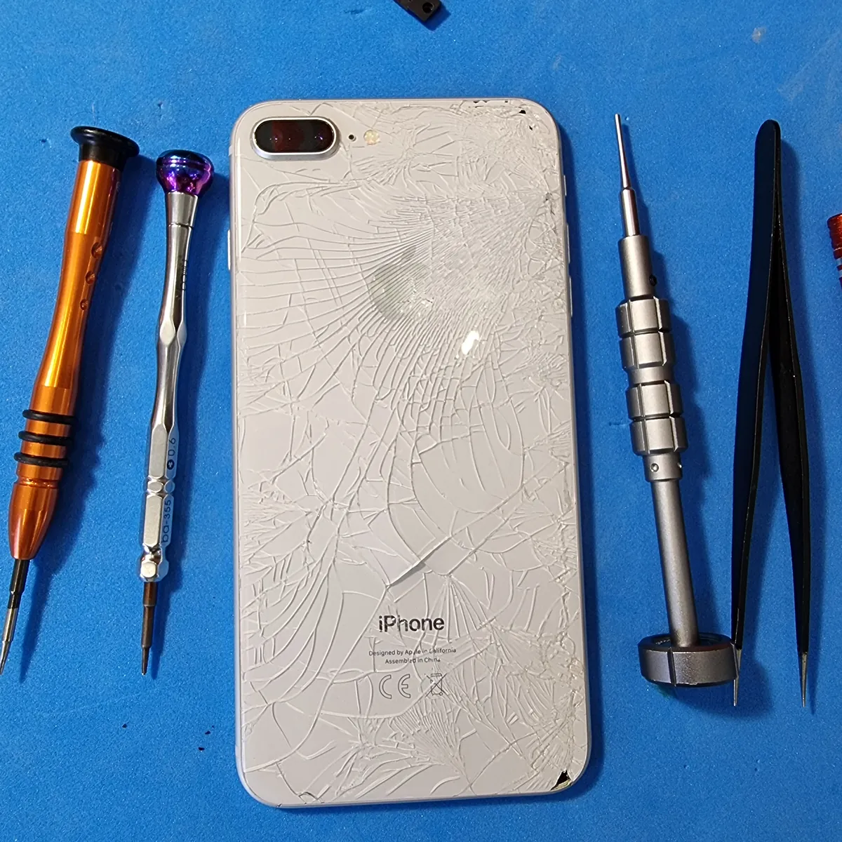 iPhone 8 Plus с разбитой в паутину задней крышкой лежит на синем ремонтном коврике между пинцетом и отвёртками мастера