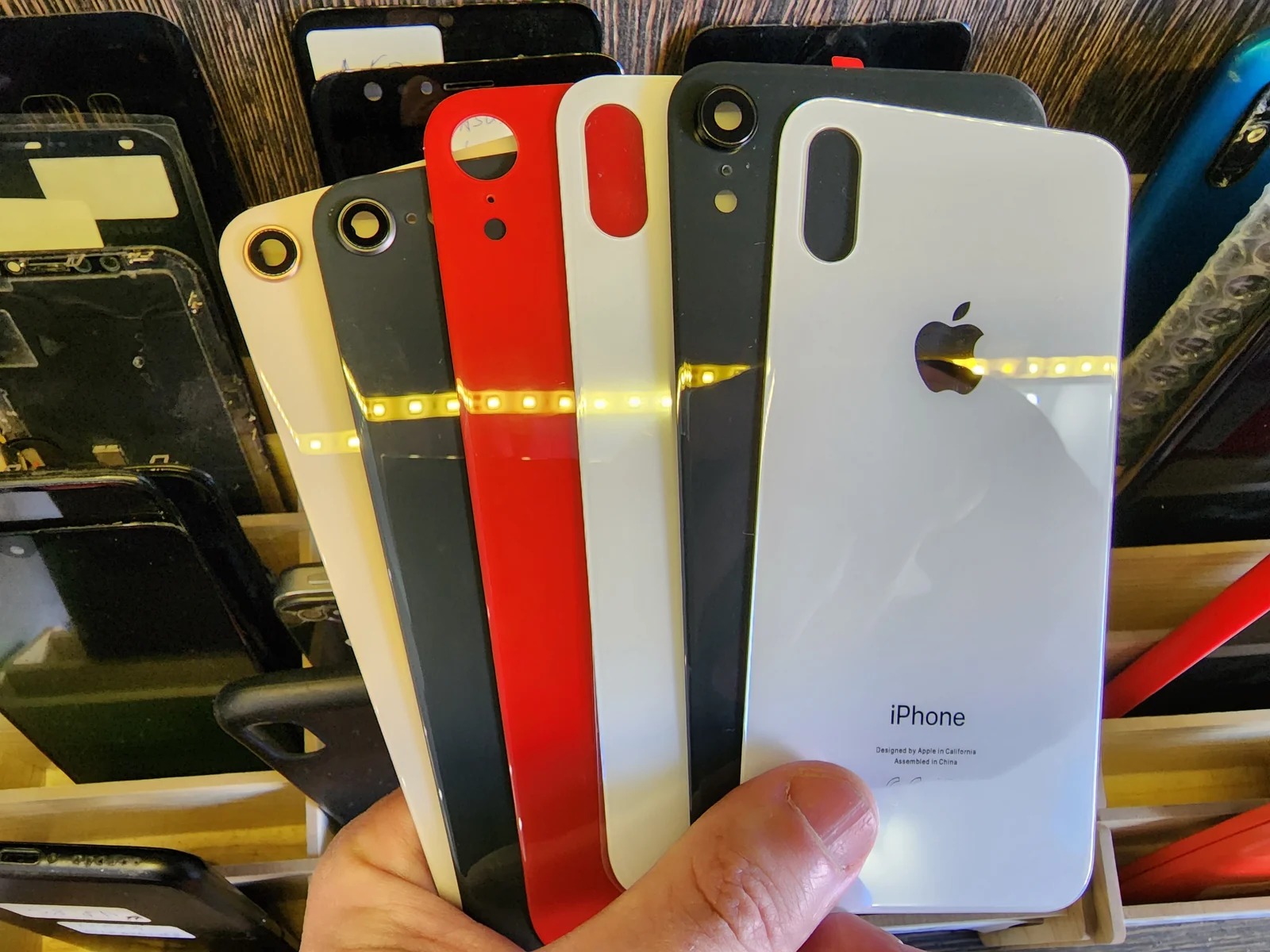Мастер держит веером шесть новых задних стёкол для iPhone разных цветов: белое, красное, чёрное, серое и бежевое, на заднем плане ящик с разобранными корпусами