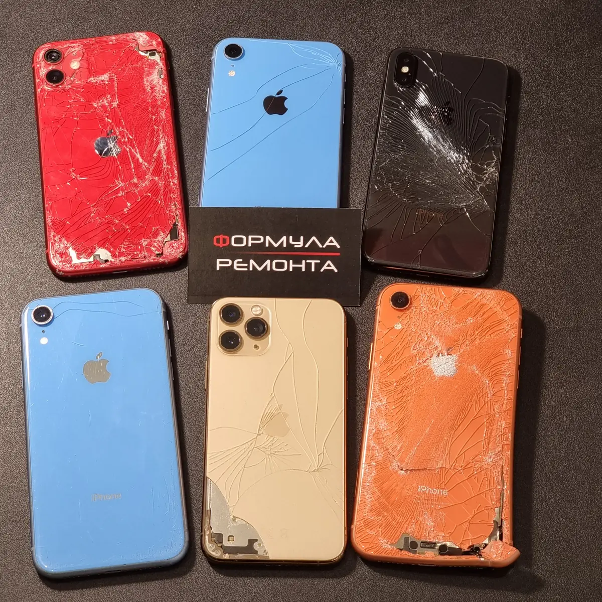 Шесть iPhone разных цветов с разбитыми задними крышками лежат на тёмном ремонтном коврике мастерской, в центре визитка сервисного центра «Формула Ремонта»