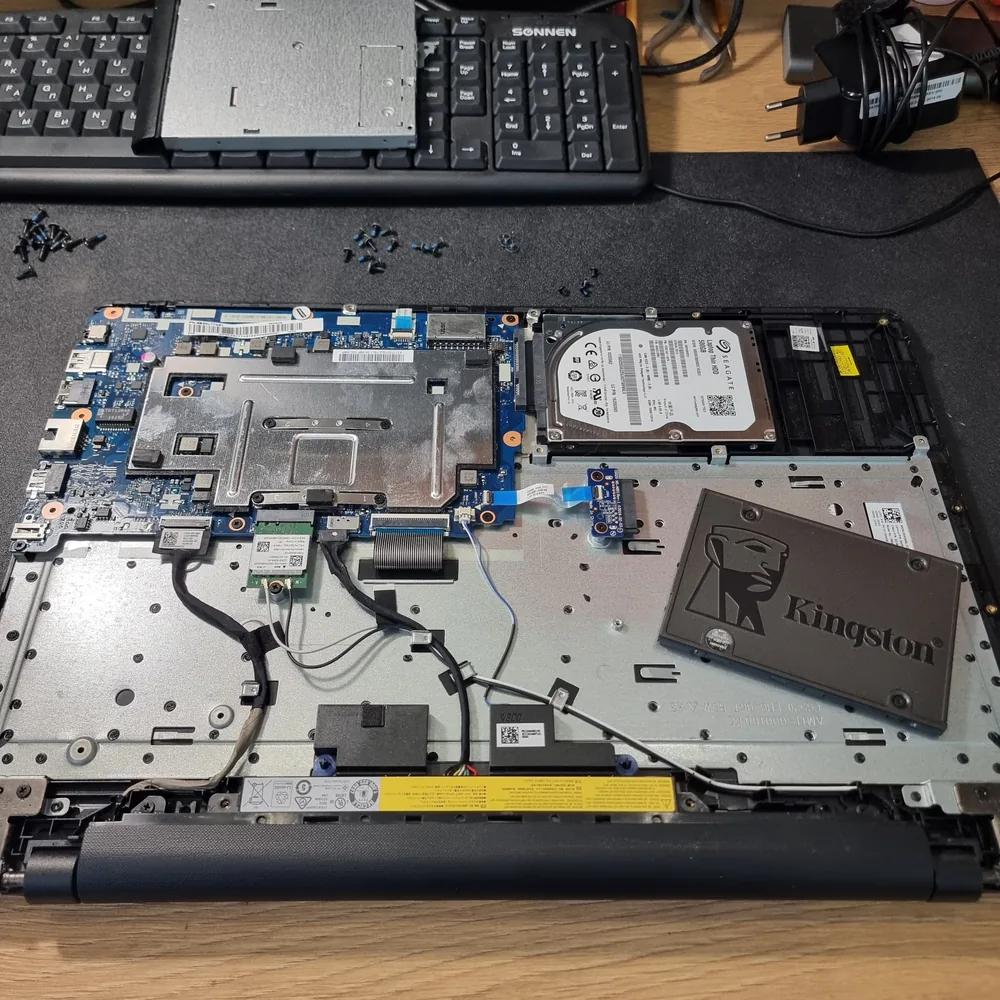 Разобранный ноутбук на столе мастерской: из отсека вынут жёсткий диск Seagate Laptop Thin на 500 гигабайт, рядом на корпусе лежит твердотельный накопитель Kingston, внизу видна батарея и снятые винты