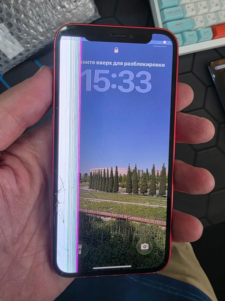 iPhone 12 mini в руке мастера: вдоль левого края экрана широкая полоса цветных вертикальных линий и трещина, остальная часть экрана показывает обои и часы