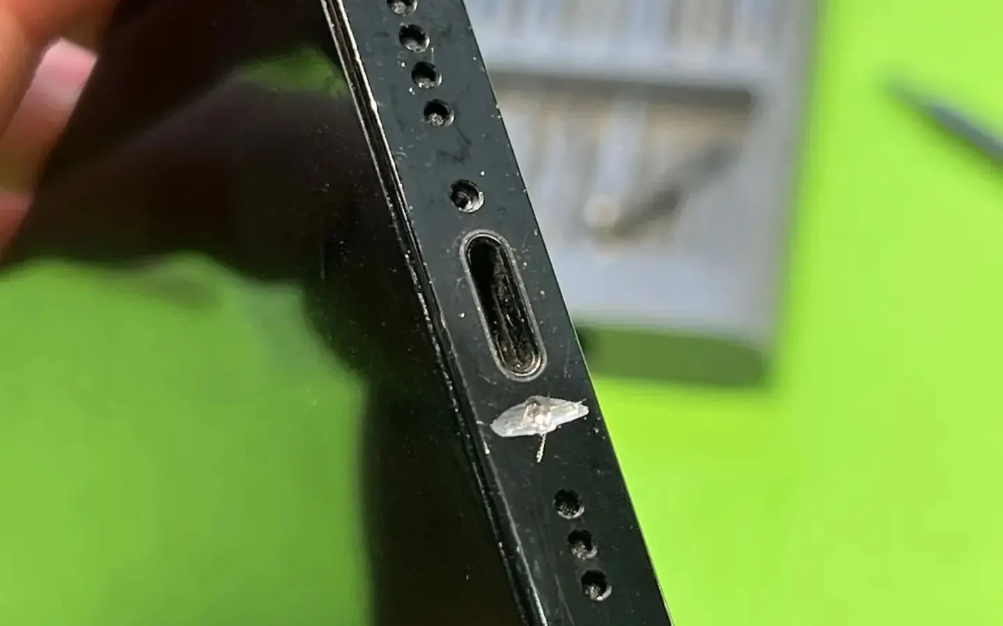 Чёрный iPhone в пальцах повёрнут нижним торцом к камере: в центре гнездо, по бокам решётки динамика и микрофона, сзади на зелёном коврике лежит набор отвёрток.