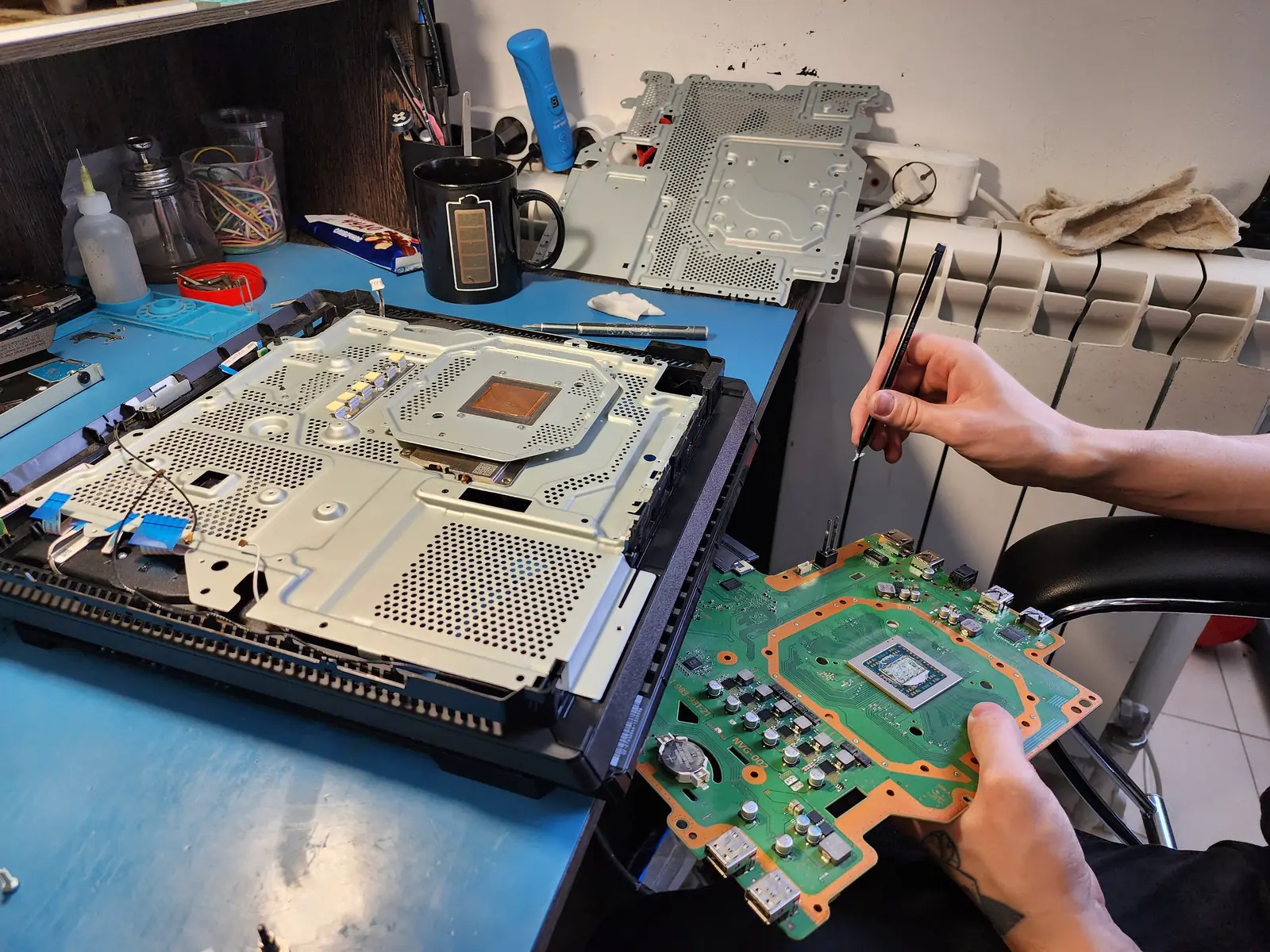Мастер держит извлечённую материнскую плату PlayStation 4 с процессором и HDMI-портом, рядом на столе лежит разобранный корпус приставки со снятым металлическим экраном