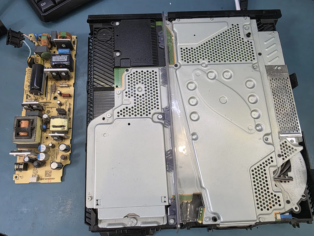 Вскрытая PS4 с металлическим экраном над платой лежит на синем рабочем коврике, в углу корпуса виден вентилятор, а слева отдельно лежит вынутая плата блока питания с конденсаторами и трансформатором