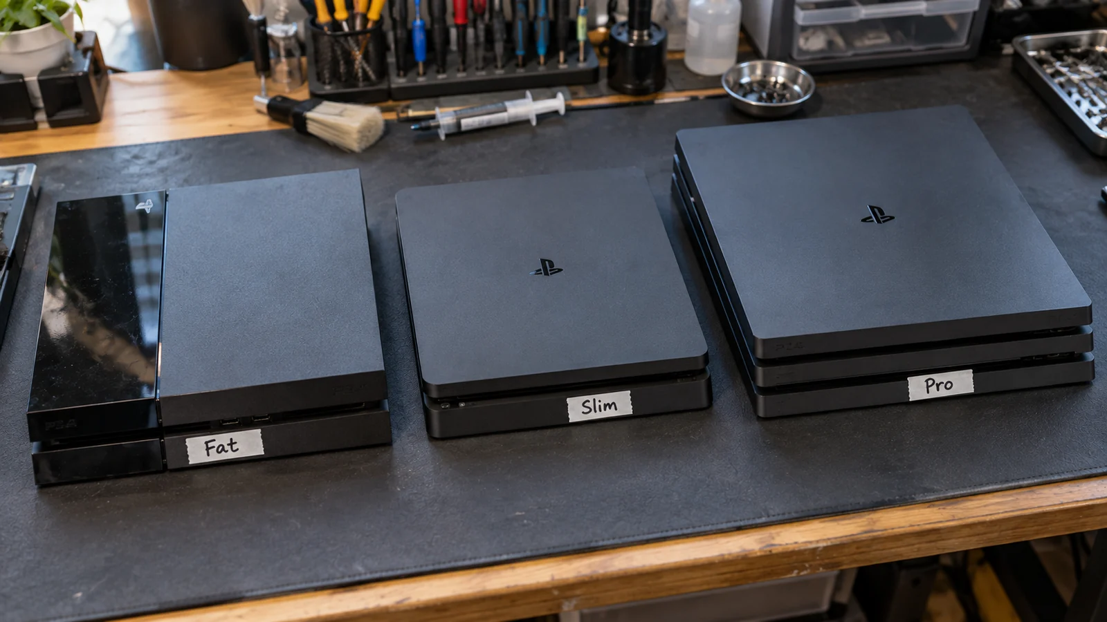 Три PlayStation 4 лежат рядом на тёмном рабочем коврике мастерской: первая PS4 с двухцветной крышкой, компактная PS4 Slim со скруглёнными краями и толстая трёхъярусная PS4 Pro, у каждой к торцу приклеена бумажная бирка с названием ревизии