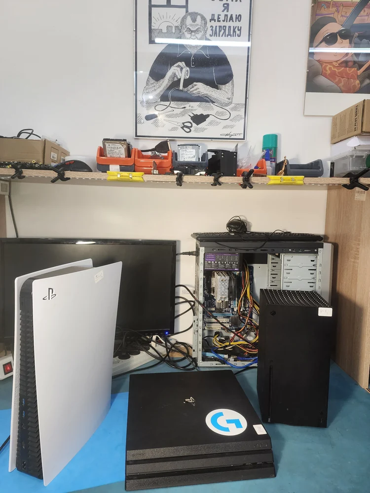 PlayStation 5 в белом корпусе стоит вертикально на столе сервисного центра, рядом лежит чёрная PS4 Pro и стоит Xbox Series X, за ними системный блок со снятой крышкой и монитор.