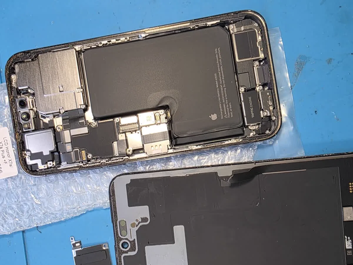 Разобранный iPhone 14 Pro Max на синем коврике мастера: в корпусе видна аккумуляторная батарея с маркировкой Apple, рядом лежит снятый дисплейный модуль