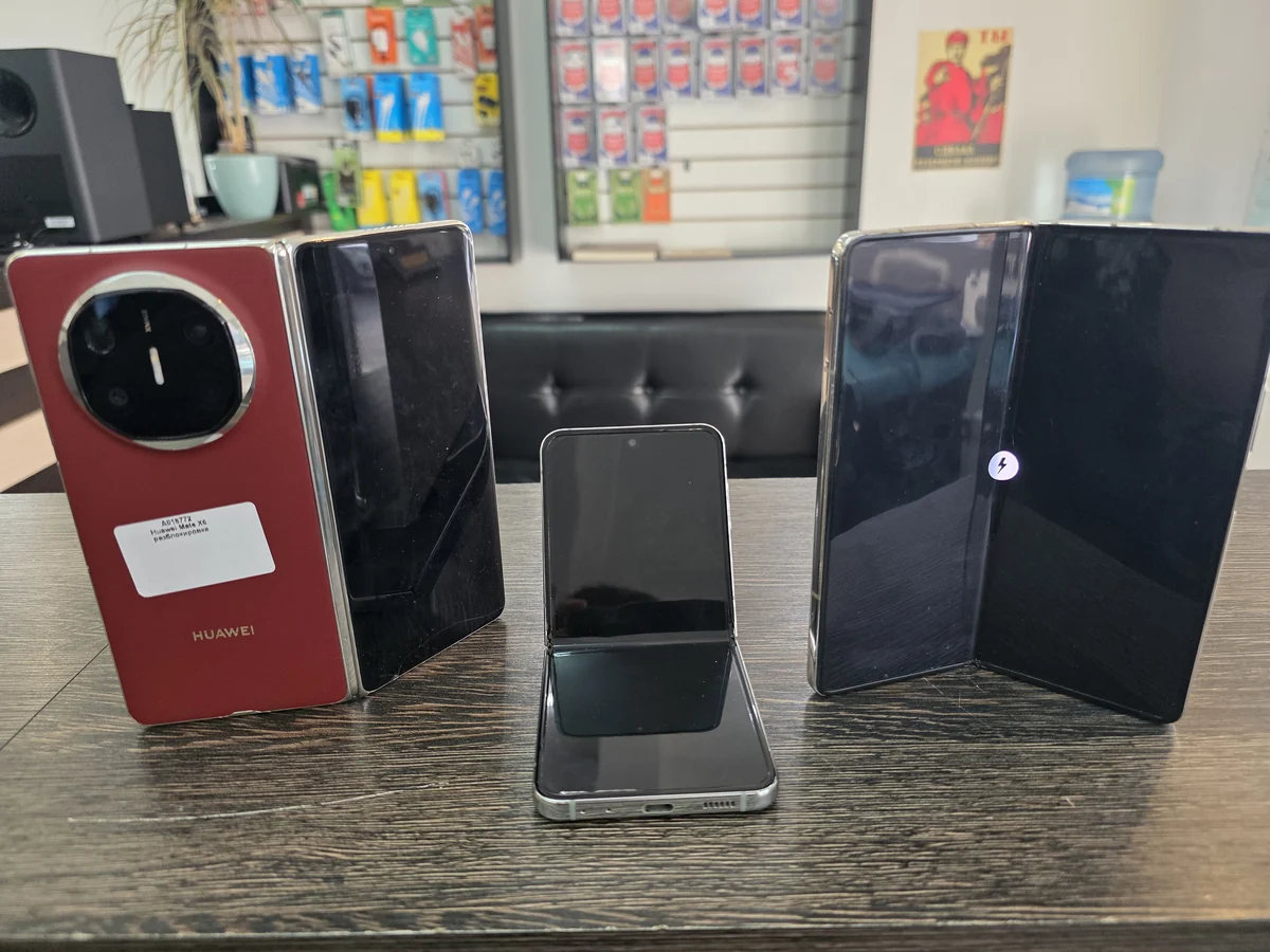На стойке приёмки мастерской три складных телефона: красный Huawei Mate X6 с белой наклейкой заказа, маленький раскрытый раскладной и большой раскрытый складной, за ними стенд с чехлами и зарядками
