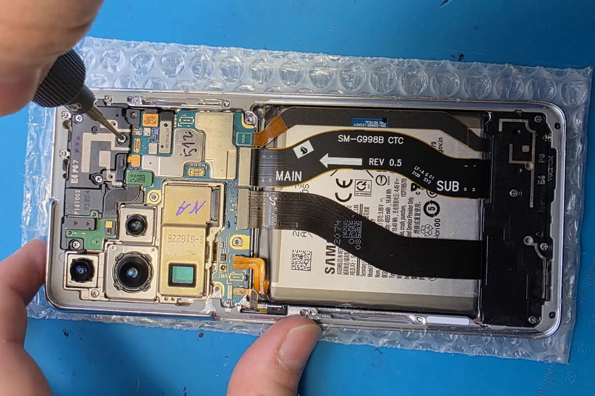 Разобранный Samsung Galaxy S21 Ultra на рабочем коврике: видны системная плата, аккумулятор и камеры, рука мастера закручивает винт отвёрткой