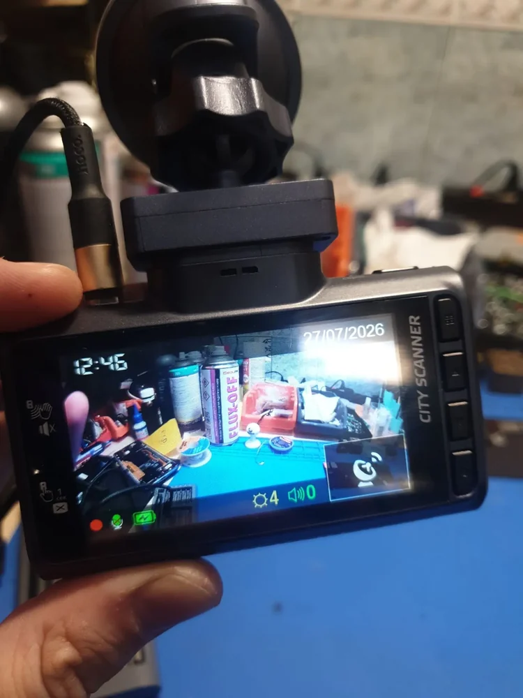 Видеорегистратор CITY SCANNER на присоске в руке над рабочим столом: к нему подключён кабель, на экране идёт картинка стола с баллончиком флюса и коробками, внизу горит красная точка.