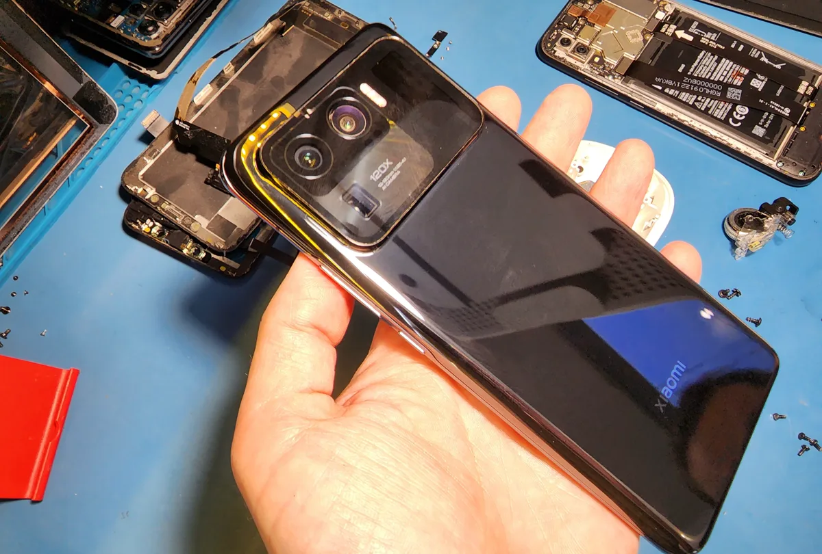 Смартфон Xiaomi в руке мастера со снятой задней крышкой, на столе рядом лежат другие разобранные аппараты и мелкие детали