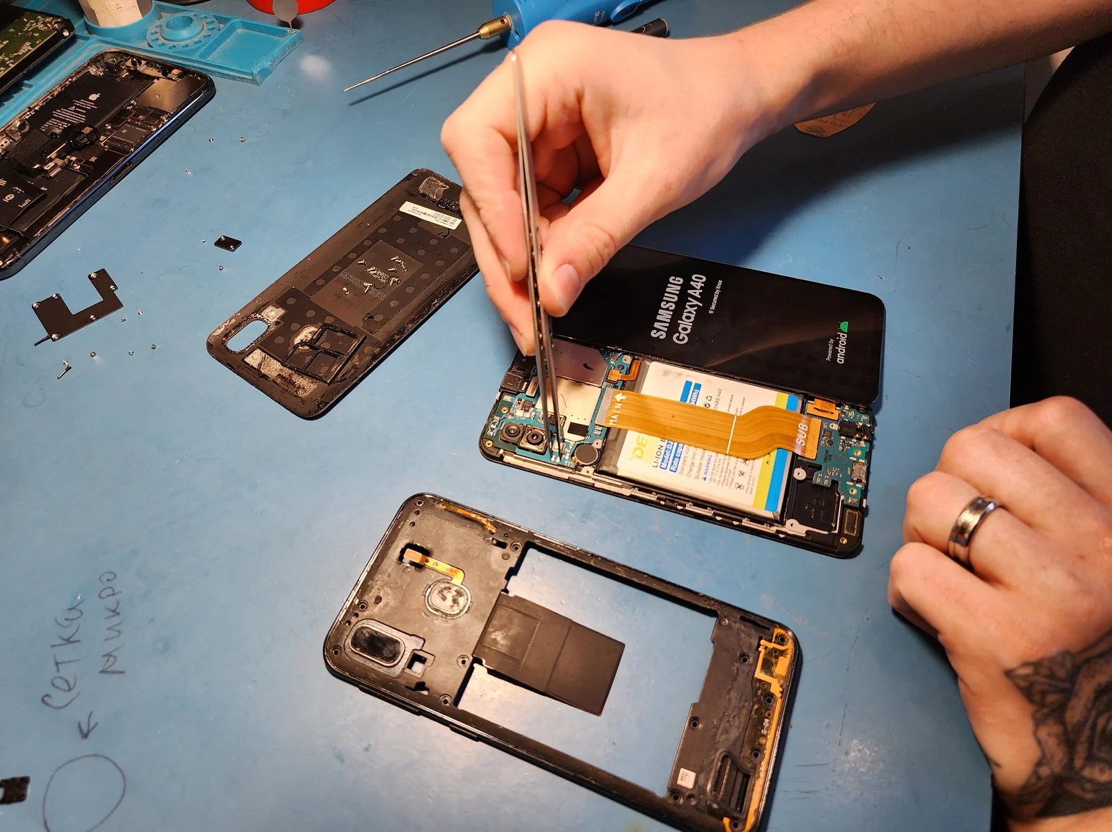 Смартфон Samsung Galaxy A40 лежит на синем рабочем коврике со снятой задней крышкой: видны аккумулятор, оранжевый шлейф и пинцет в руке мастера.