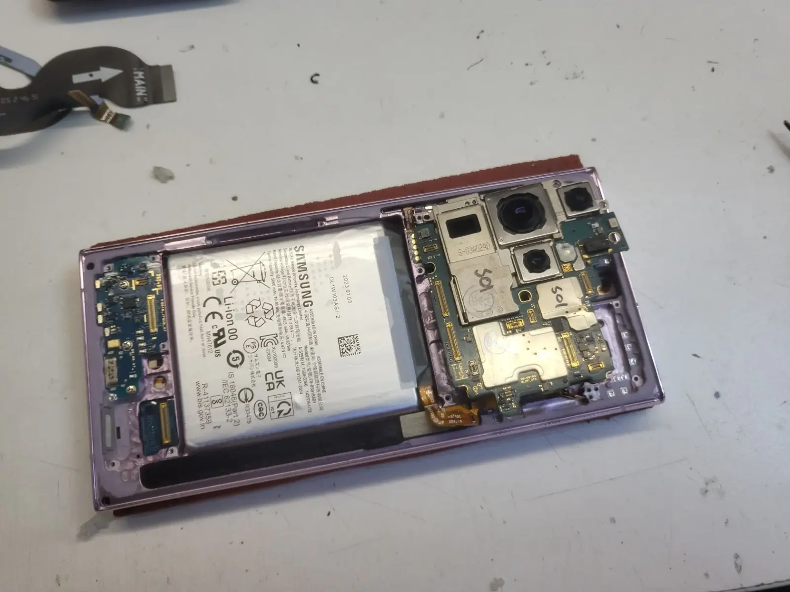 Вскрытый Samsung Galaxy S23 Ultra в лавандовом корпусе лежит на рабочем столе: снята задняя крышка, видны аккумулятор, основная плата и блок камер.