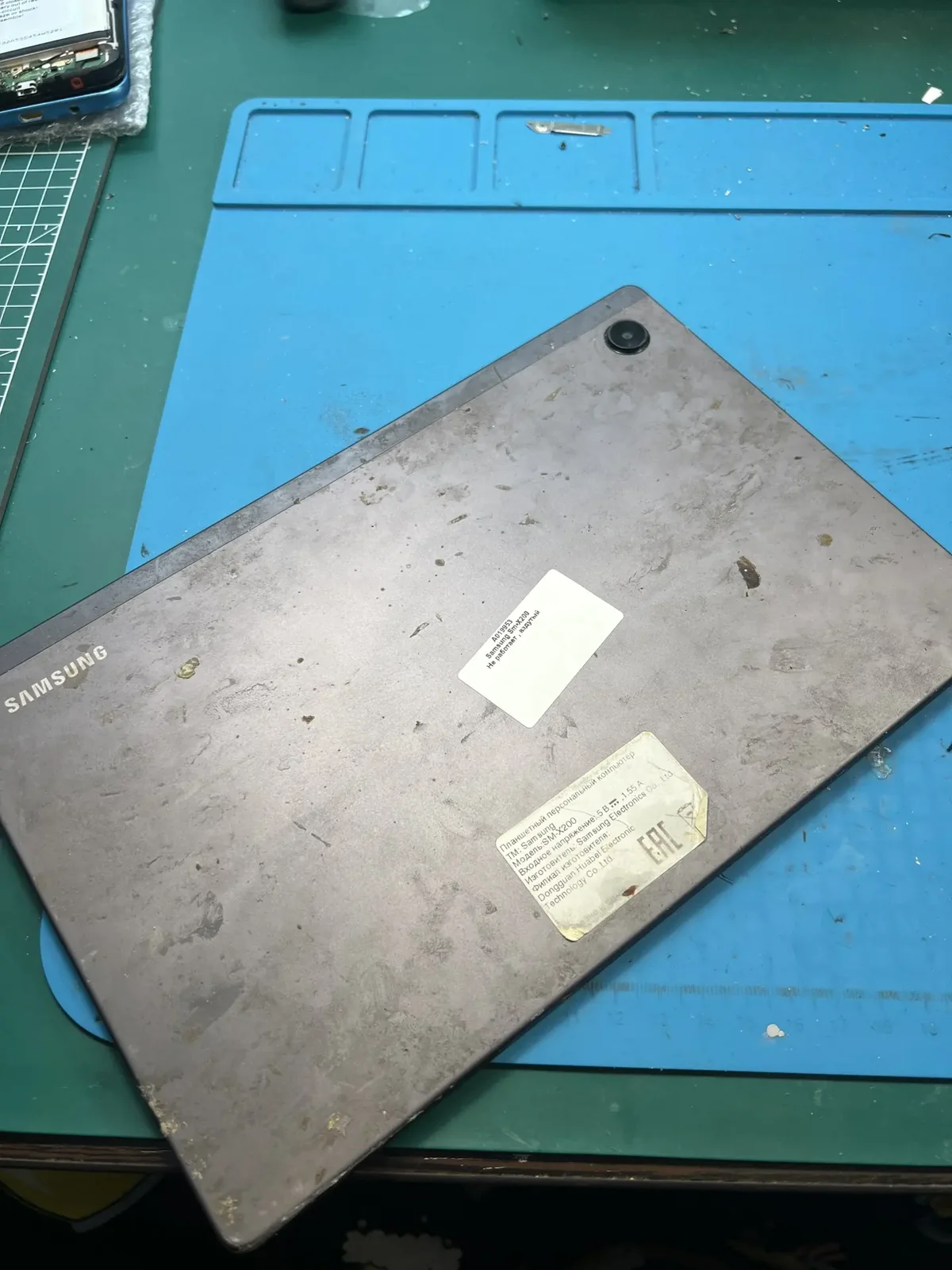 Планшет Samsung Galaxy Tab A8 с наклейками на задней крышке лежит на синем ремонтном коврике перед разбором