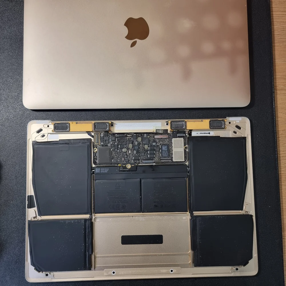 Открытый MacBook золотистого цвета лежит на чёрном коврике: секции аккумулятора лежат в нижней половине корпуса слева, справа и посередине, вокруг места трекпада.
