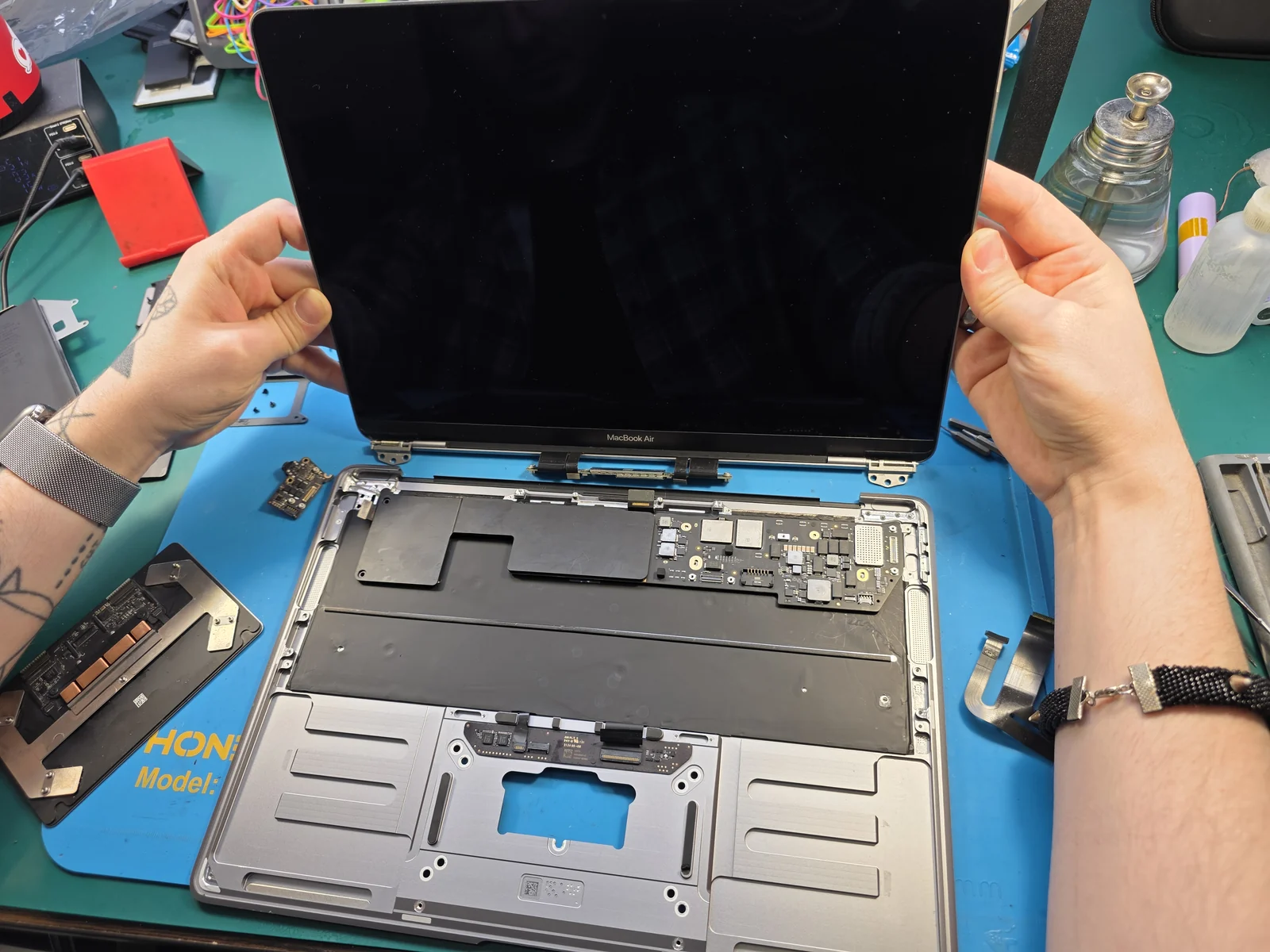 Разобранный MacBook Air на синем коврике, мастер поднял крышку с экраном: трекпад снят и лежит слева, в корпусе на его месте видна прямоугольная прорезь и плата с разъёмами шлейфов.