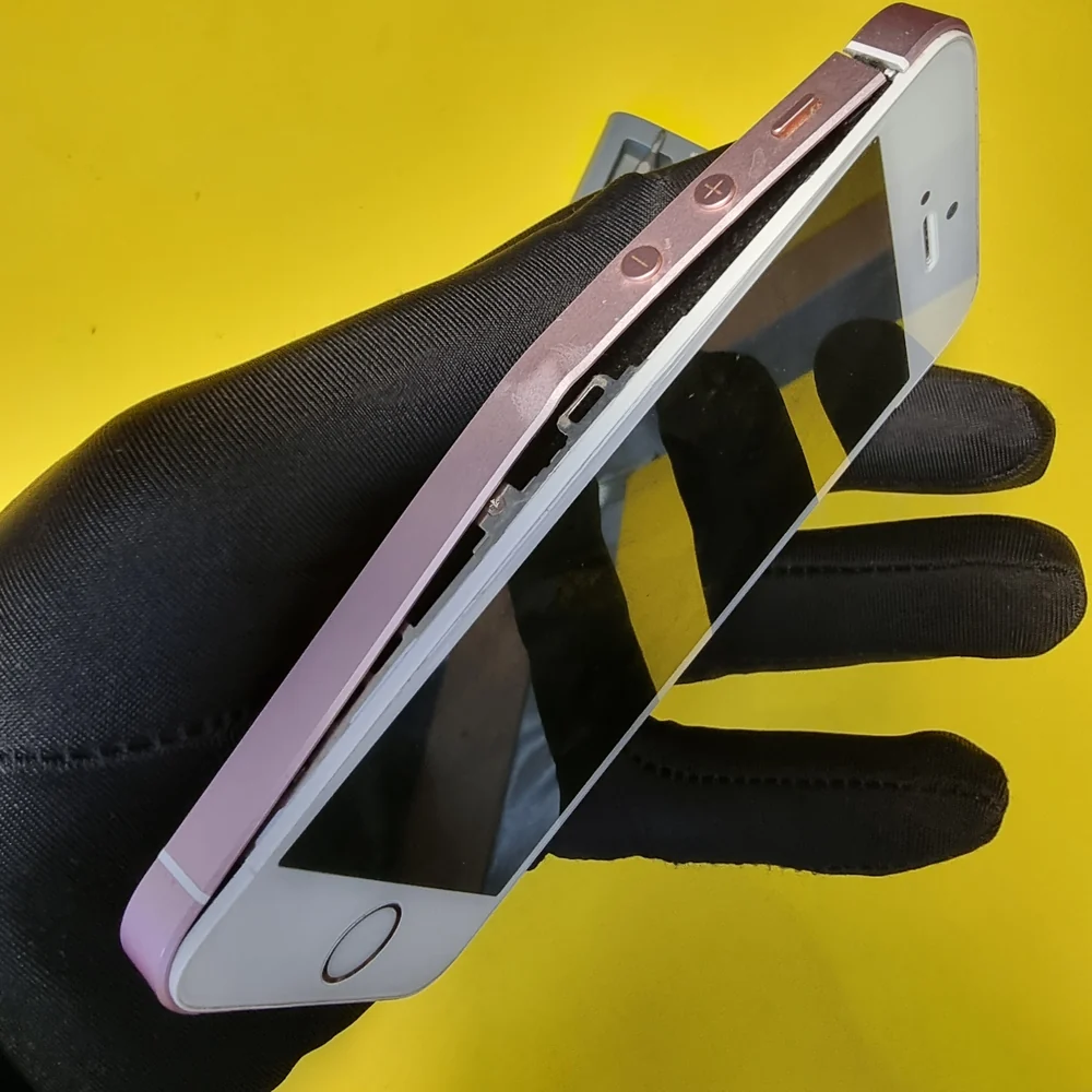 iPhone SE с розовым корпусом в руке мастера в чёрной перчатке: вздувшийся аккумулятор приподнял экран, в щели между экраном и корпусом видны внутренние детали.