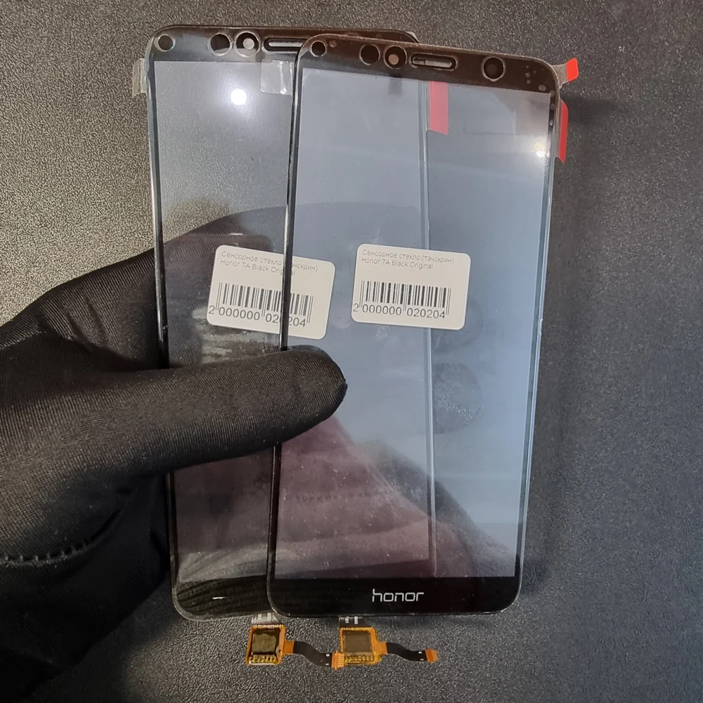 В руке мастера в чёрной перчатке два новых сенсорных стекла для Honor 7A с наклейками-штрихкодами, снизу свисают оранжевые шлейфы.
