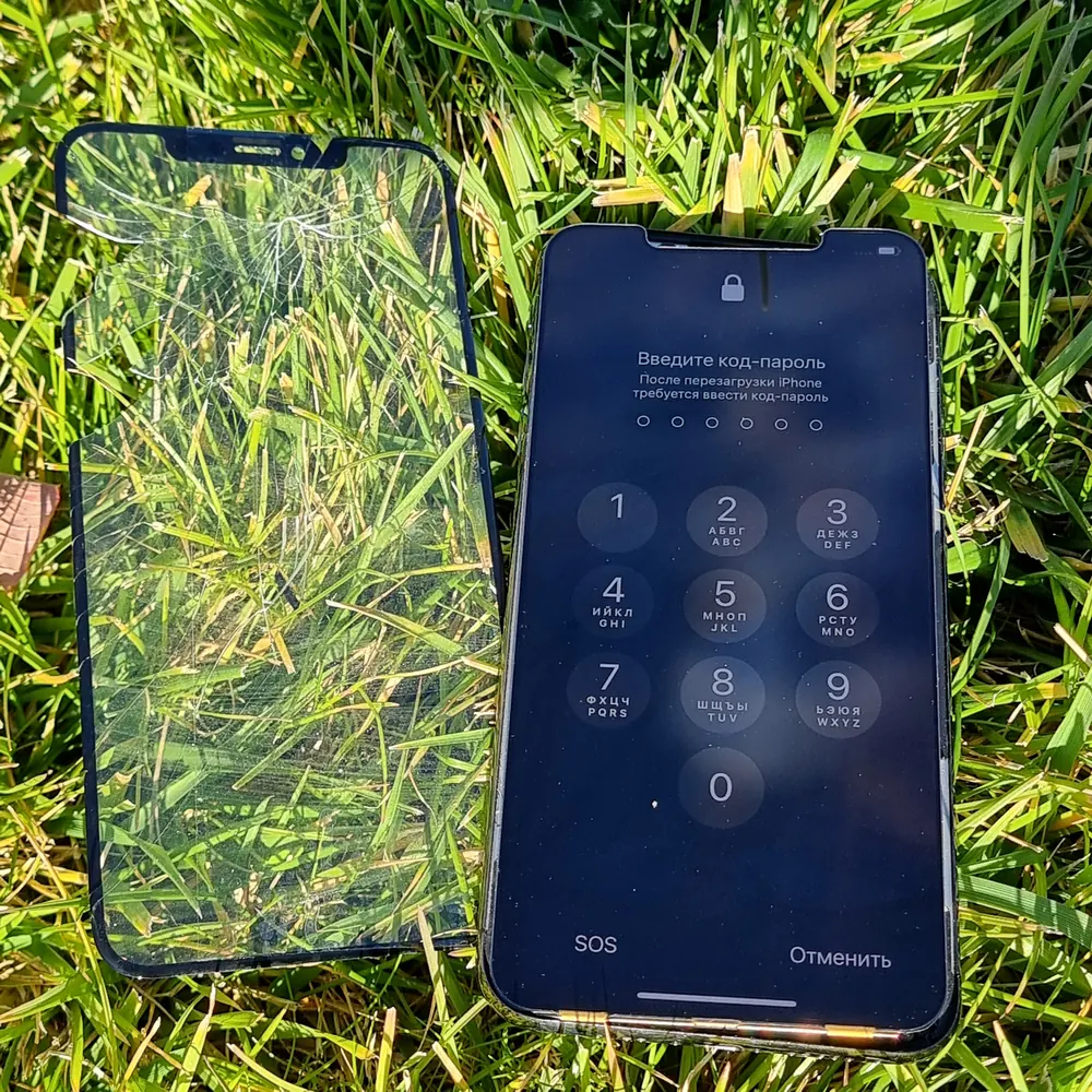 iPhone лежит на траве и просит ввести код-пароль, рядом лежит снятое защитное стекло, всё в трещинах.