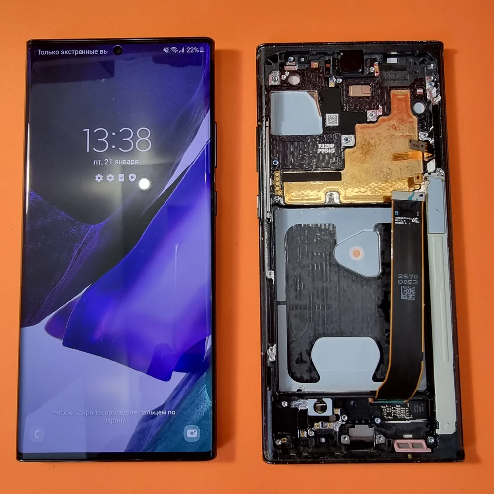 Samsung Galaxy Note 20 Ultra в разборе на оранжевом столе: слева снятый дисплей, который продолжает работать и показывает время, справа рамка с платой и шлейфами