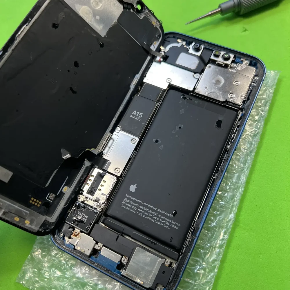 Вскрытый iPhone 13 на зелёном коврике мастерской: на изнанке дисплея и на аккумуляторе видны капли воды, рядом лежит отвёртка.