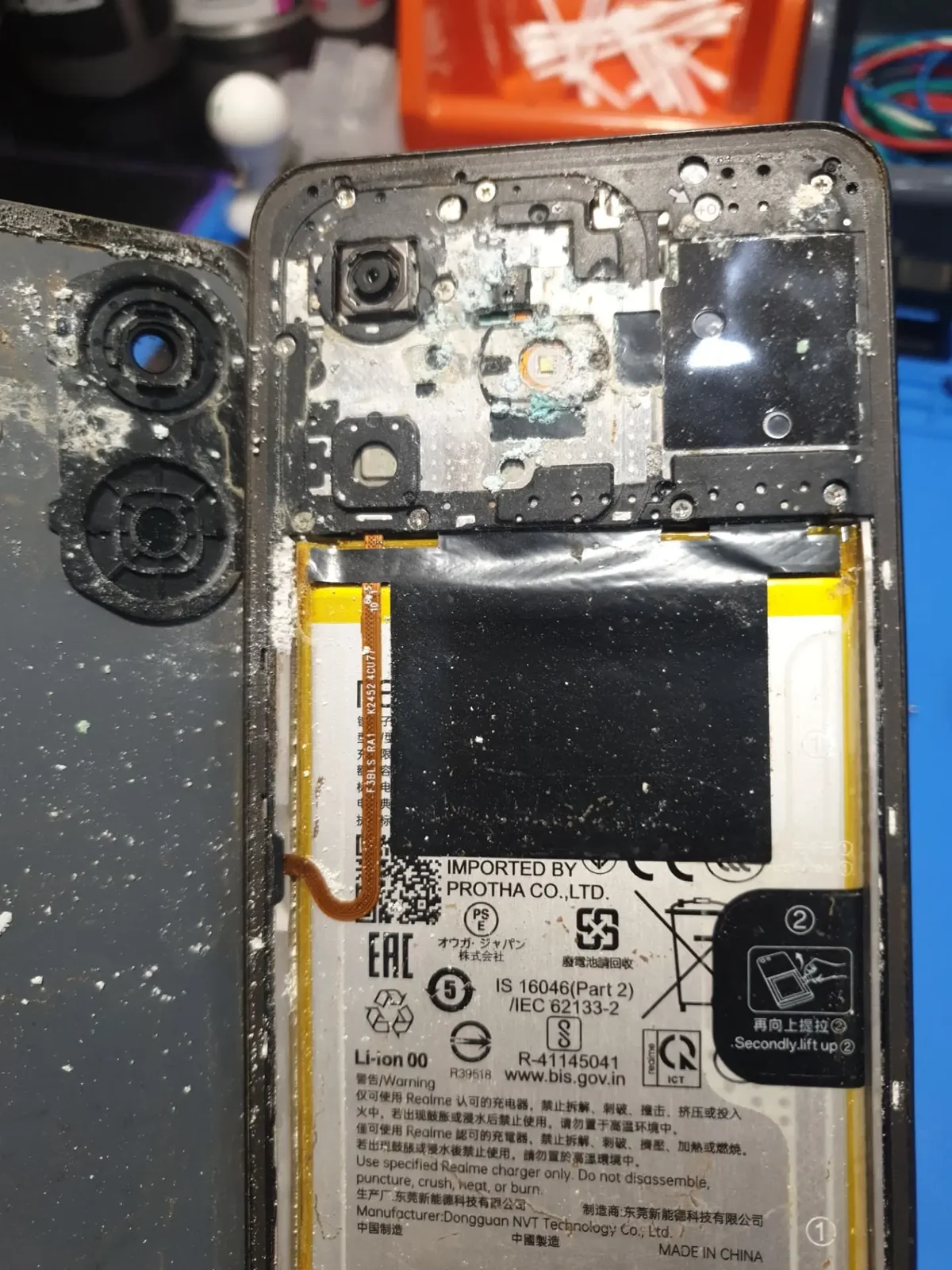 Смартфон Realme со снятой задней крышкой на столе мастера: по периметру корпуса белый налёт высохших солей, рядом со вспышкой бирюзовое пятно окисления