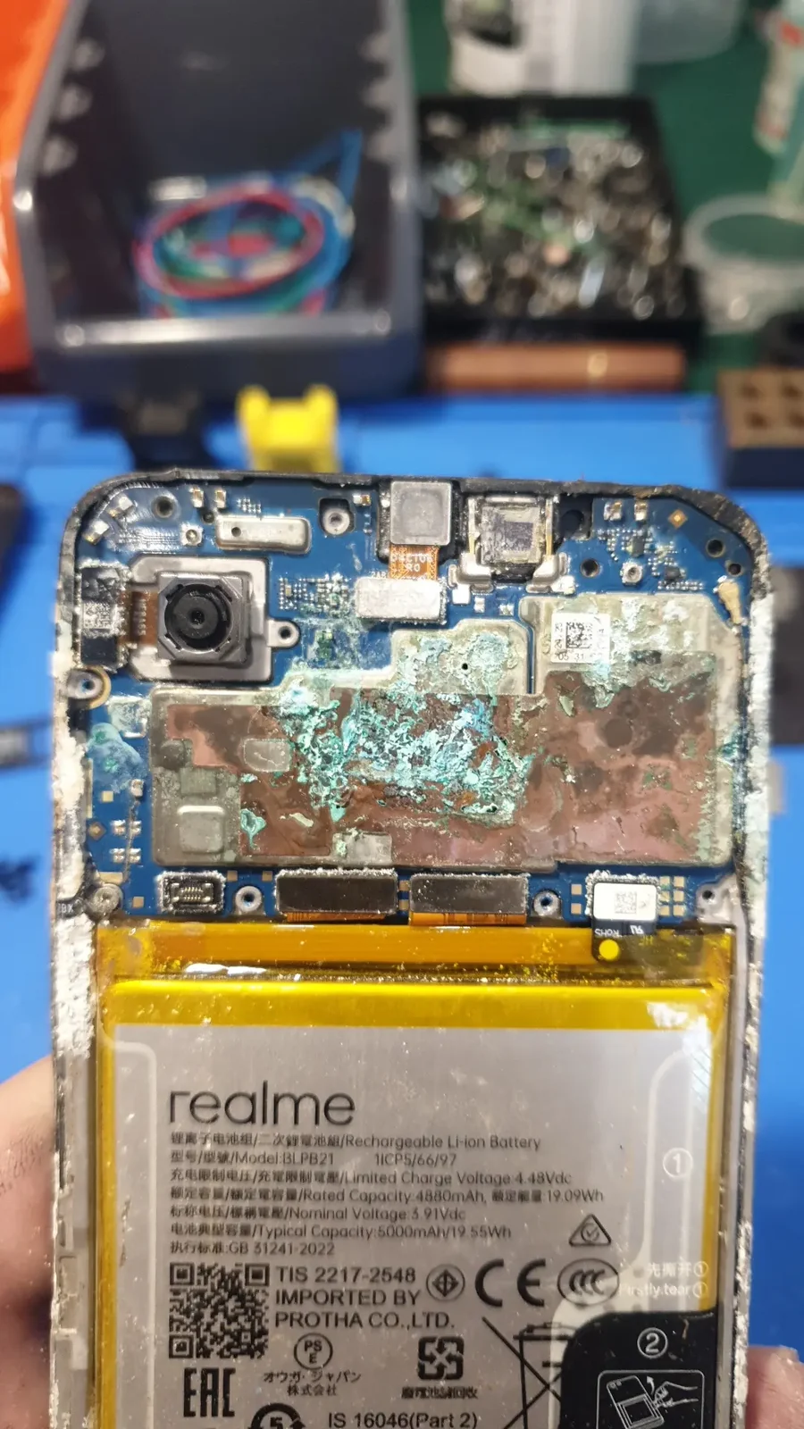 Плата смартфона Realme крупным планом: центральная часть покрыта бирюзовыми разводами окислившейся меди, по краям белый солевой налёт, снизу видна маркировка аккумулятора