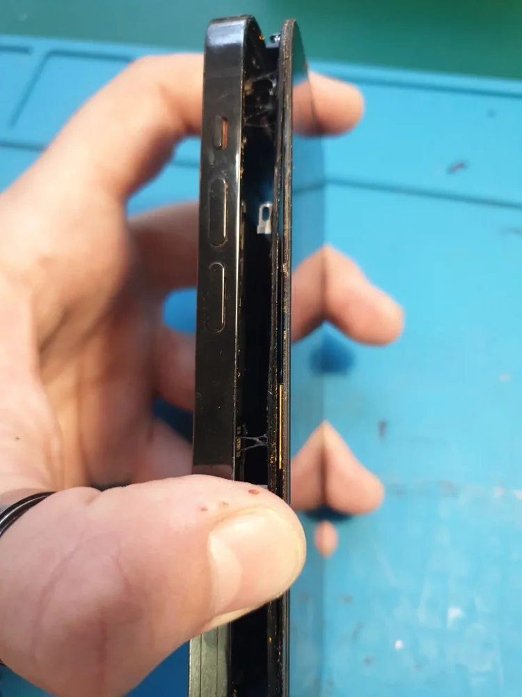 iPhone в руке мастера повёрнут торцом к камере: экран отошёл от металлической рамки, в щели видны внутренние детали, внизу синий ремонтный коврик.